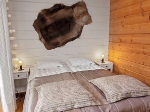 Un dormitorio con un tapete de piel grande colgando del techo, dos mesitas de noche con lámparas, una cama con colcha color beige y marrón, y paredes de paneles de madera.