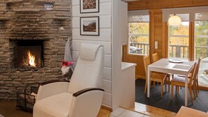 A cozy living room with a stone fireplace, a reclining chair, a dining area with a table and chairs, and a view of autumn trees through a window. - Vuosseli-kelkka 15a by Interhome (Kuusamo)