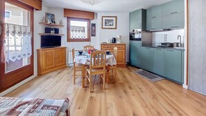 Une cuisine de taille réduite avec des meubles en bois, une table à manger avec des chaises, un téléviseur et une fenêtre avec un rideau.