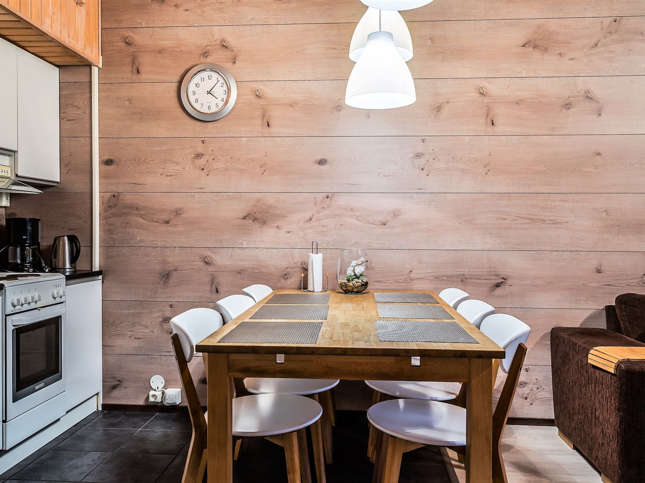 Une table de salle à manger en bois, des chaises blanches, une horloge au mur et une cuisine moderne équipée d’une cuisinière et d’une machine à café.