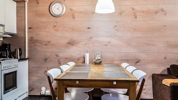 A wooden dining table with white chairs, a clock on the wall, and a modern kitchen with a stove and coffee maker.