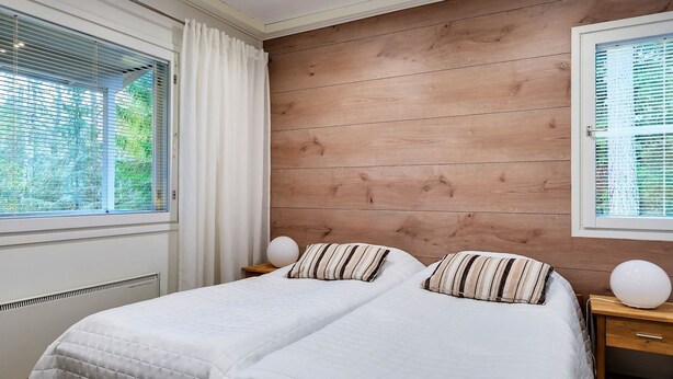 A bedroom with a large bed, two bedside tables, a wooden wall panel, and a window with curtains. - Weekend family by Interhome (Sotkamo)