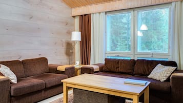 A cozy living room with two sofas, a coffee table, and a lamp. There are large windows with blinds, a wooden ceiling, and a rug on the floor.