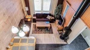 A cozy cabin interior with a wooden floor, a sofa, a coffee table, a dining table with chairs, a fireplace, and a large window with curtains. - Weekend family by Interhome (Sotkamo)