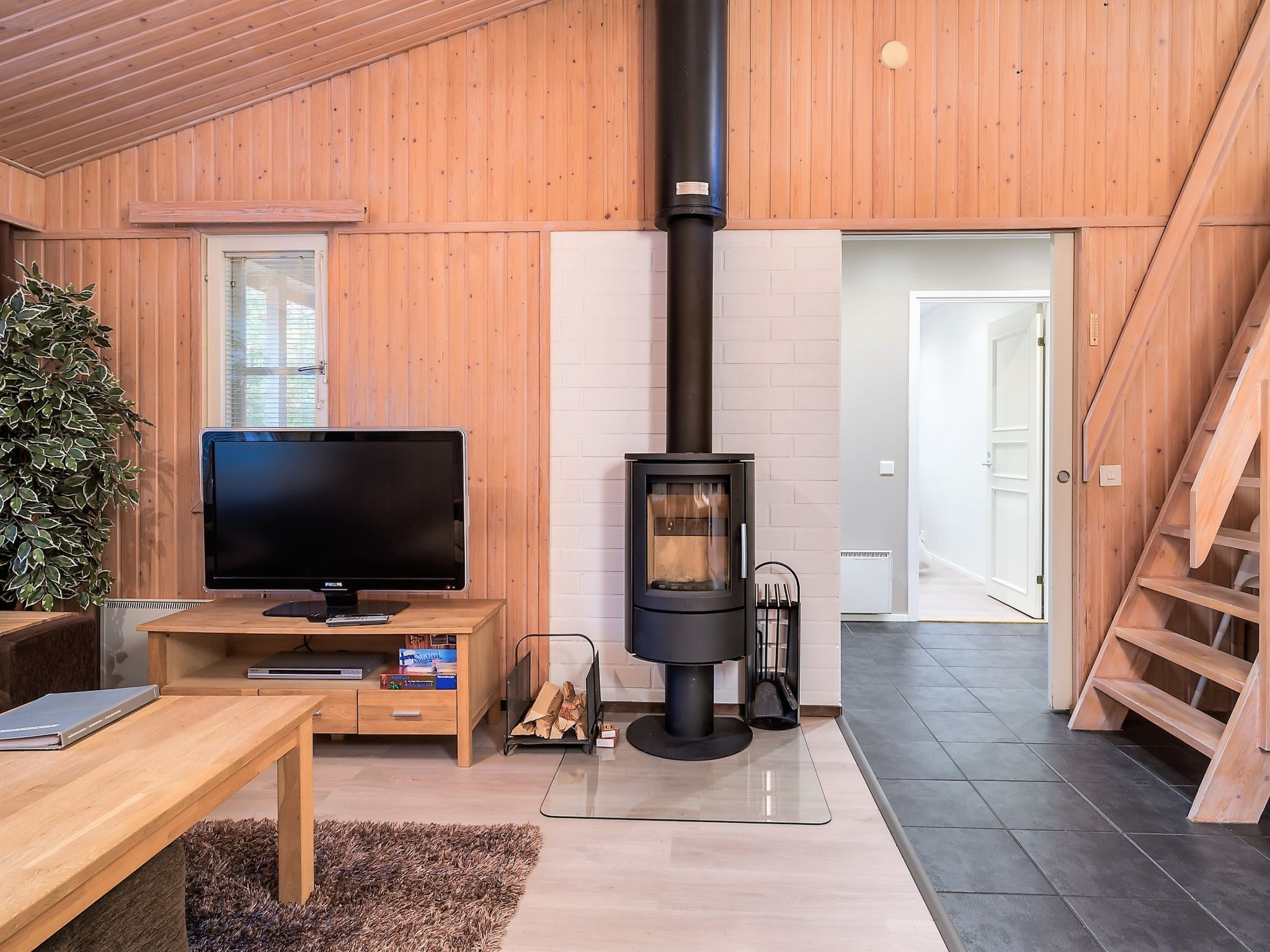 A modern living room with a large flat-screen TV, a wooden TV stand, a black wood-burning stove, and a staircase.