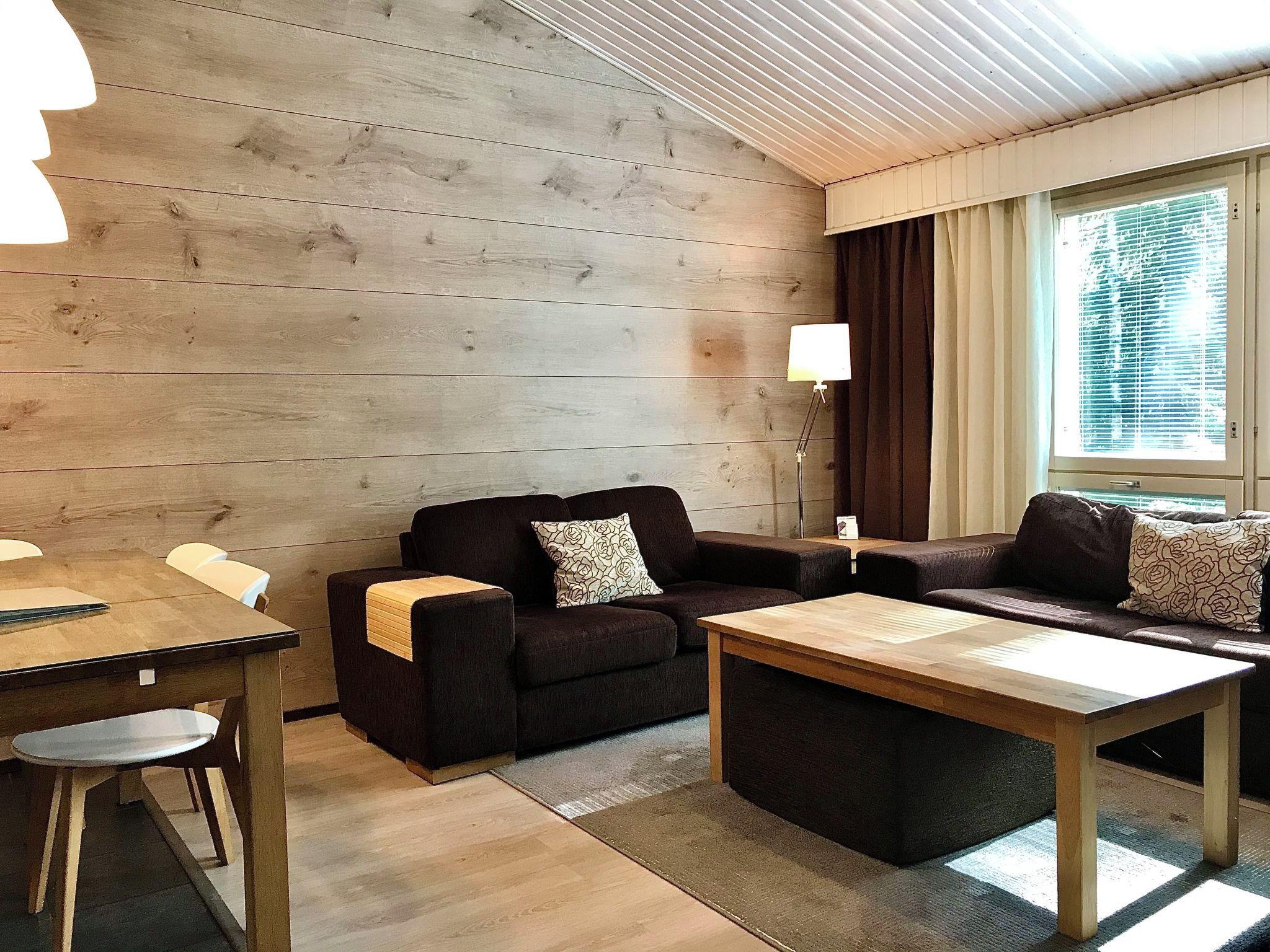 A cozy living room with a wooden wall, a sofa set, a coffee table, and a dining table with chairs.