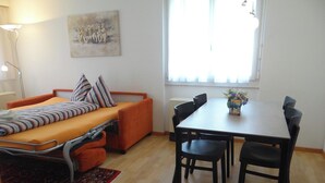 Una habitación pequeña con cama, una mesa y sillas, una ventana con cortinas y una lámpara.