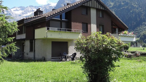 Una casa de dos pisos con balcón, rodeada por un patio cubierto de césped y montañas al fondo.