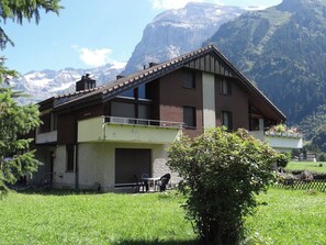 Una casa de dos pisos con balcón, rodeada por un patio cubierto de césped y montañas al fondo.