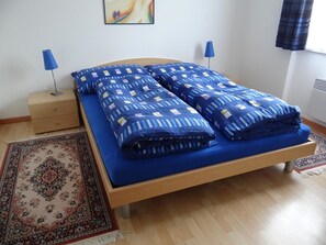 Una cama con una colcha y almohadas azules, una mesita de noche con una lámpara y una alfombra estampada en el suelo.