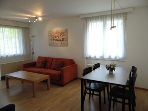 A living room with a red sofa, a wooden coffee table, a dining table with chairs, and a painting on the wall.