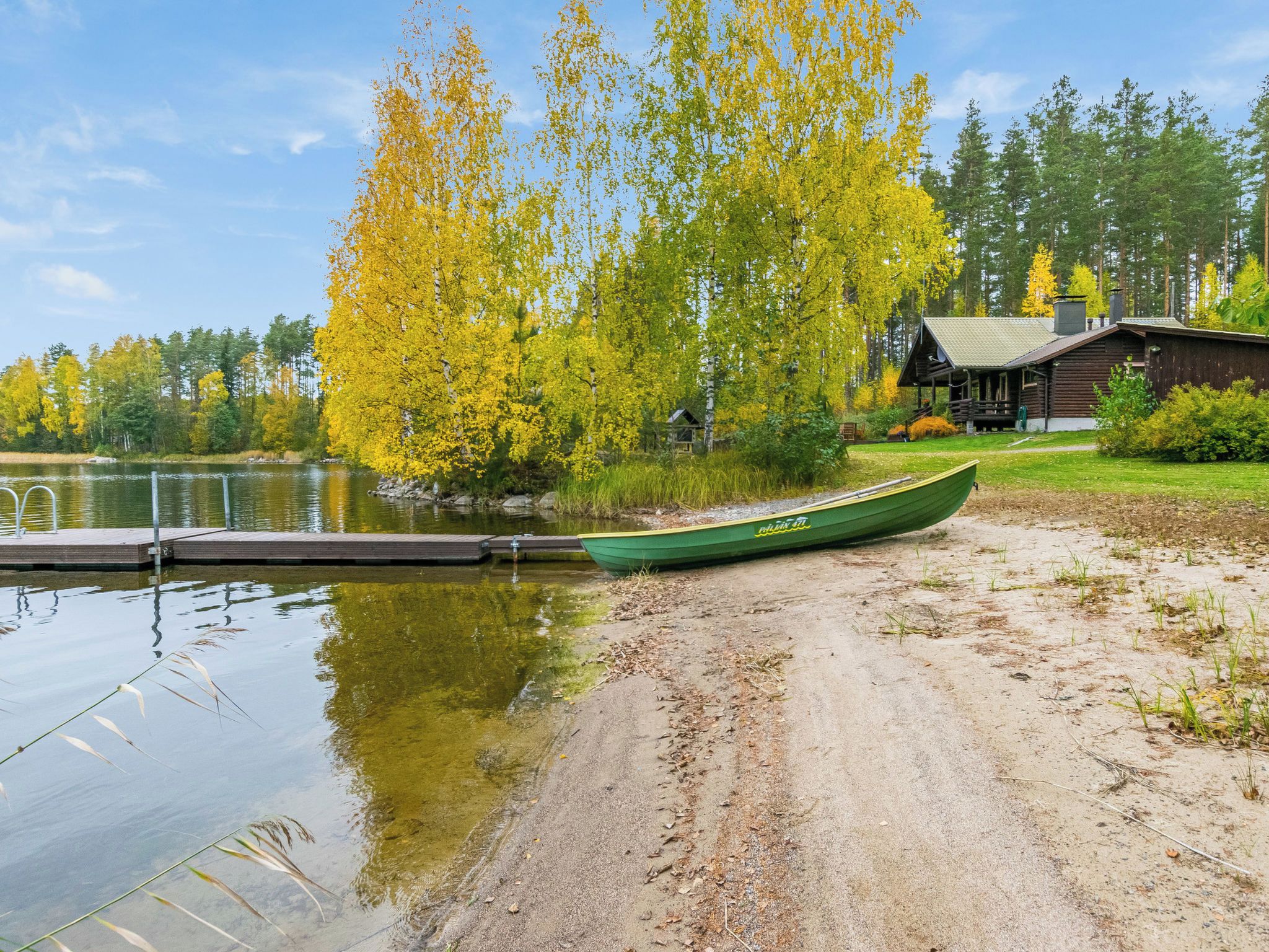 A green canoe on a sandy shore by a calm lake, with a wooden cabin and trees in the background.