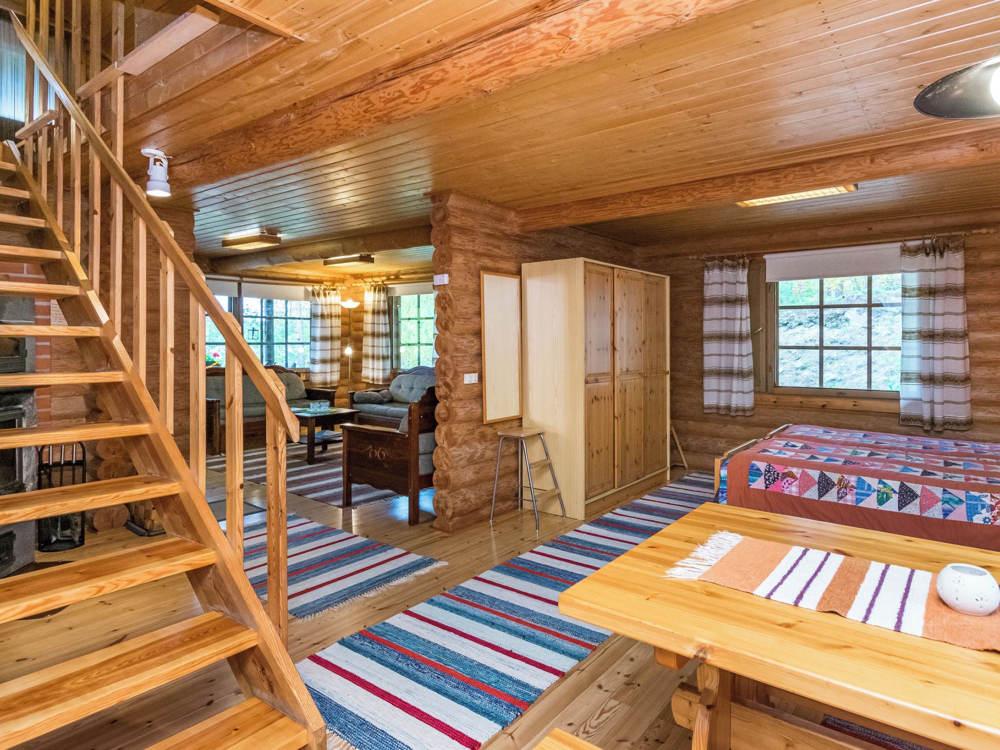 L'intérieur d'une cabane en rondins, avec un escalier en bois, une cheminée, un canapé, un lit et une fenêtre avec des rideaux.