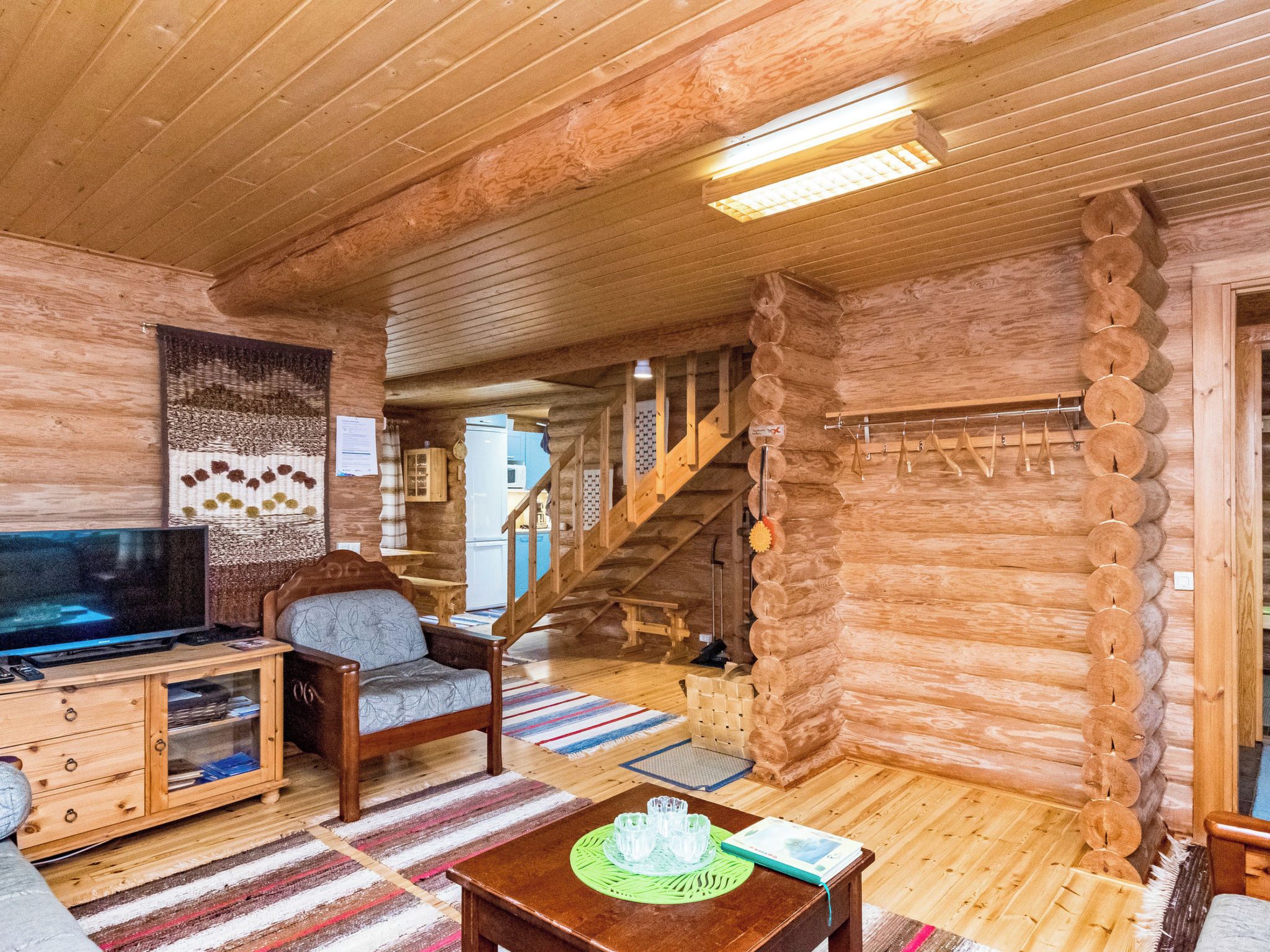 Interior de uma cabana de tronco com televisão, uma cadeira, uma mesa com copos e uma escada.