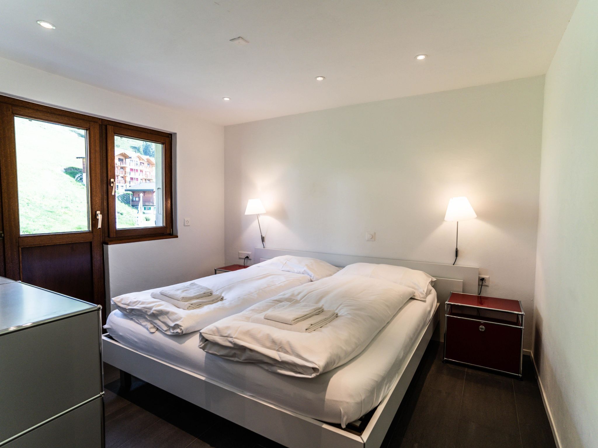 A modern hotel room with a bed, bedside lamps, a desk, and a view through a window.