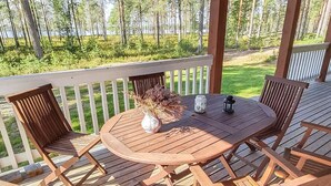 Eine Holzterrasse mit rundem Tisch und Stühlen, umgeben von einem weißen Geländer, mit Blick auf ein bewaldetes Gebiet.