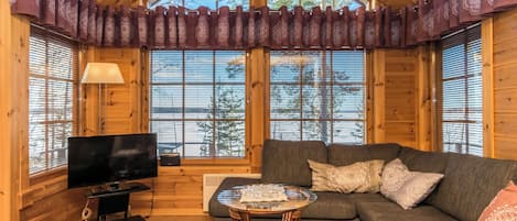 A cozy cabin interior with a wooden ceiling, large windows, a television, a sofa, and a small table.