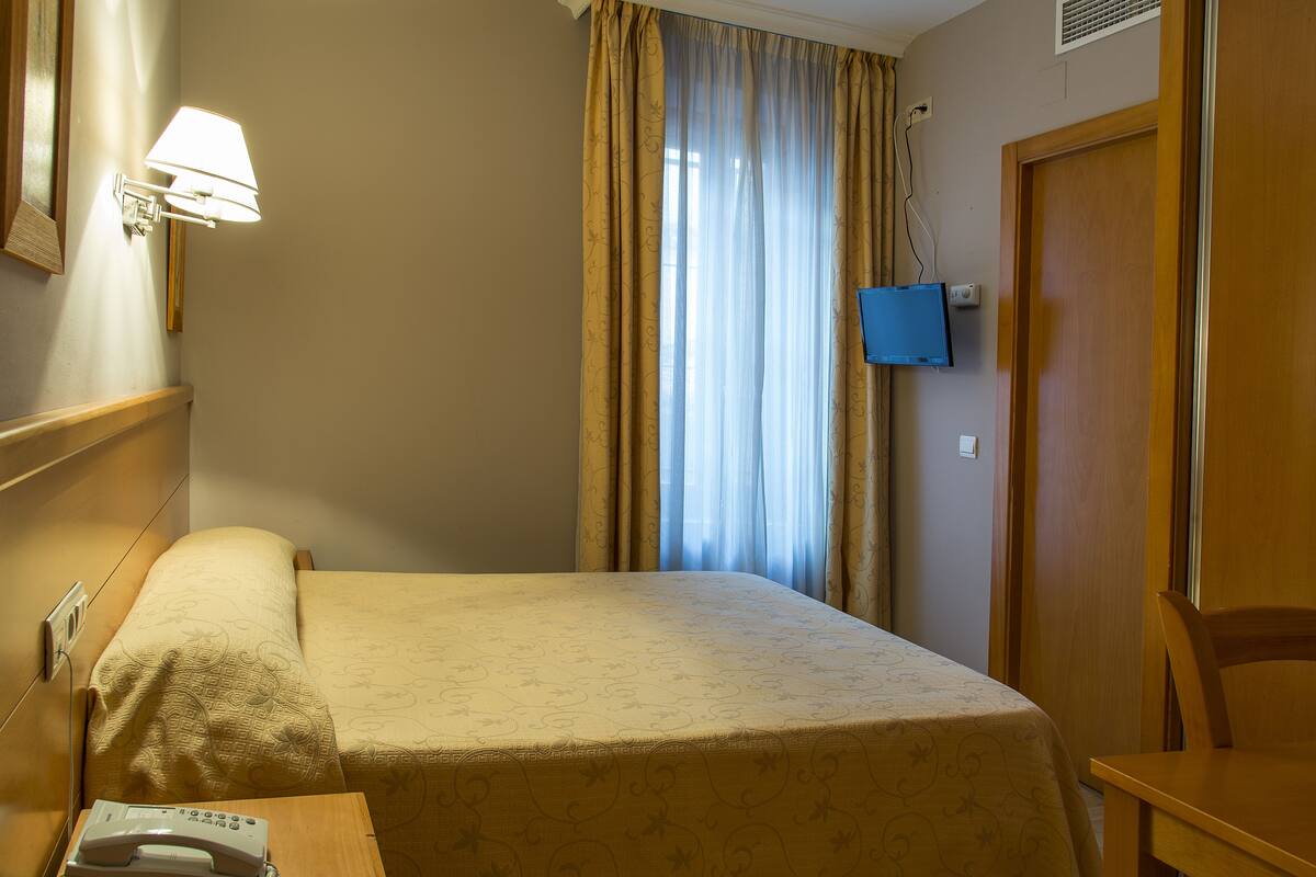 Una habitación de hotel con cama, mesita de noche, lámpara, ventana con cortinas y televisión.