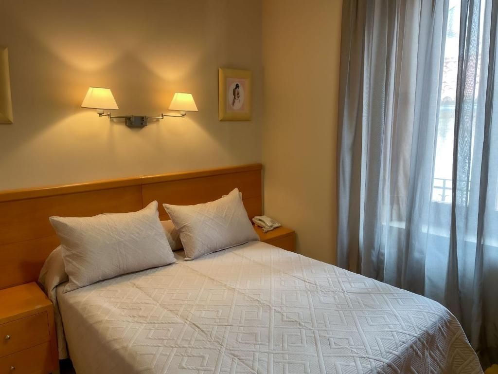 Habitació d'hotel amb un llit, dues làmpades de nit, una taula de nit, un telèfon i una finestra amb cortines.