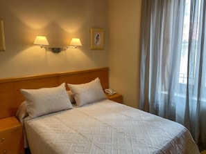 A hotel room with a bed, two bedside lamps, a nightstand, a telephone, and a window with curtains. - Hotel Castilla (Cáceres)