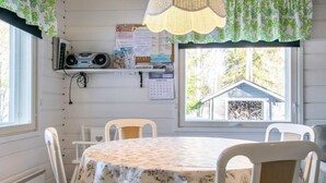 Een eetruimte met een ronde tafel, bedekt met een bloemendecor, omringd door witte stoelen. Er is een raam met uitzicht op een huis en bomen, en een radio aan de muur met verschillende documenten en een agenda.