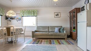 Een gezellige woonkamer met een bank, een eettafel met stoelen, een keukenhoek met een koelkast, en een raam met gordijnen.
