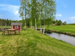 Eine rote Holzhütte mit einem kleinen Tisch und Stühlen, eine Holzbrücke über einen Bach und ein Wiesenfeld mit Birkenbäumen.