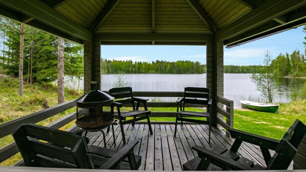Een overdekt terras buiten met een barbecue, stoelen en uitzicht op een meer, omringd door bomen.