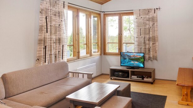 A cozy living room with a sofa, a coffee table, and a TV on a stand. There are large windows with curtains, a radiator, and a small wooden table.