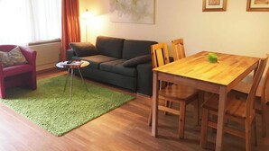 Ett vardagsrum med en soffa, ett litet bord, ett matbord med stolar, en grön matta och en tavla på väggen.