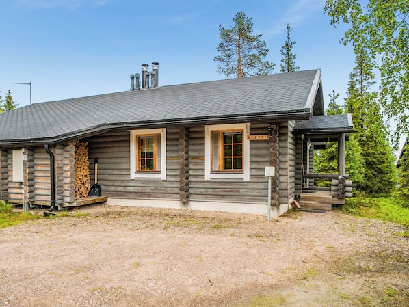 A log cabin with two windows and a chimney, surrounded by trees.