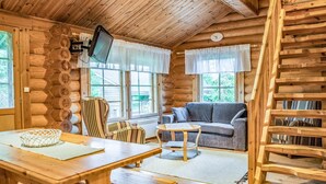 Interior acogedor de una cabaña de troncos con techo de madera, un televisor montado en la pared, un sofá gris, un sillón a rayas y una mesa de madera con una cesta.