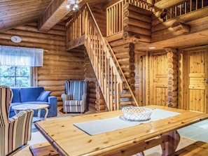 A log cabin interior with a wooden staircase, a sofa, a striped armchair, and a dining table.