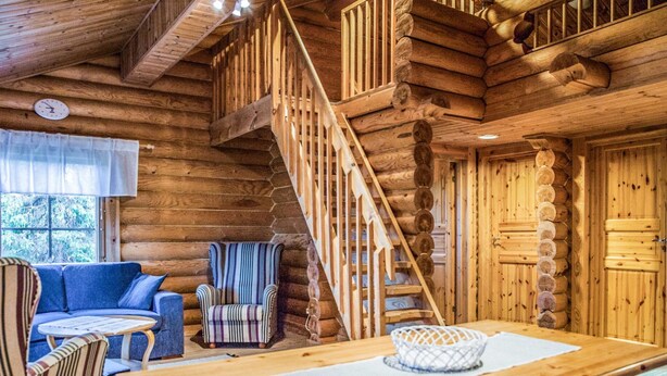 Interior de una cabaña de troncos con una escalera de madera, un sofá, un sillón a rayas y una mesa de comedor.