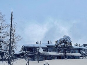 Uma cabana coberta de neve com uma chaminé grande, rodeada por árvores carregadas de neve.