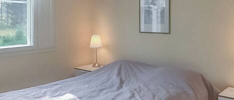 Een slaapkamer met een bed, een nachtkastje, een lamp, een raam met gordijnen en een lijst met een afbeelding aan de muur.