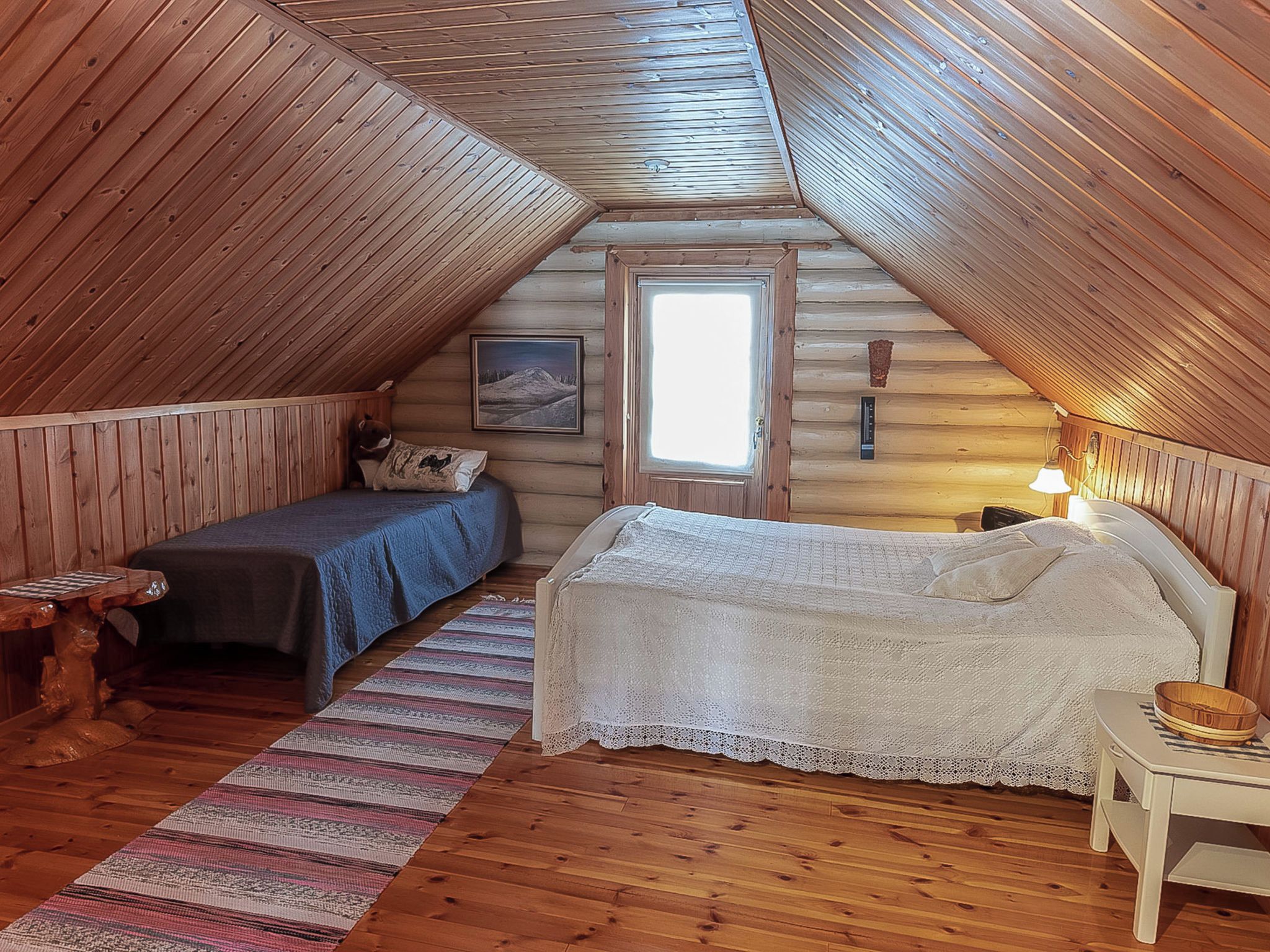 Un dormitorio acogedor en el ático, con techo inclinado, cama, banco, mesita y ventana.