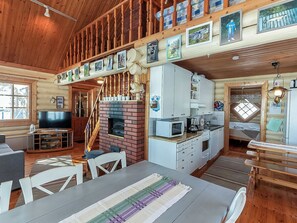 Un intérieur de chalet chaleureux avec une cheminée, un coin repas, une cuisine dotée de meubles blancs et un téléviseur dans le salon.