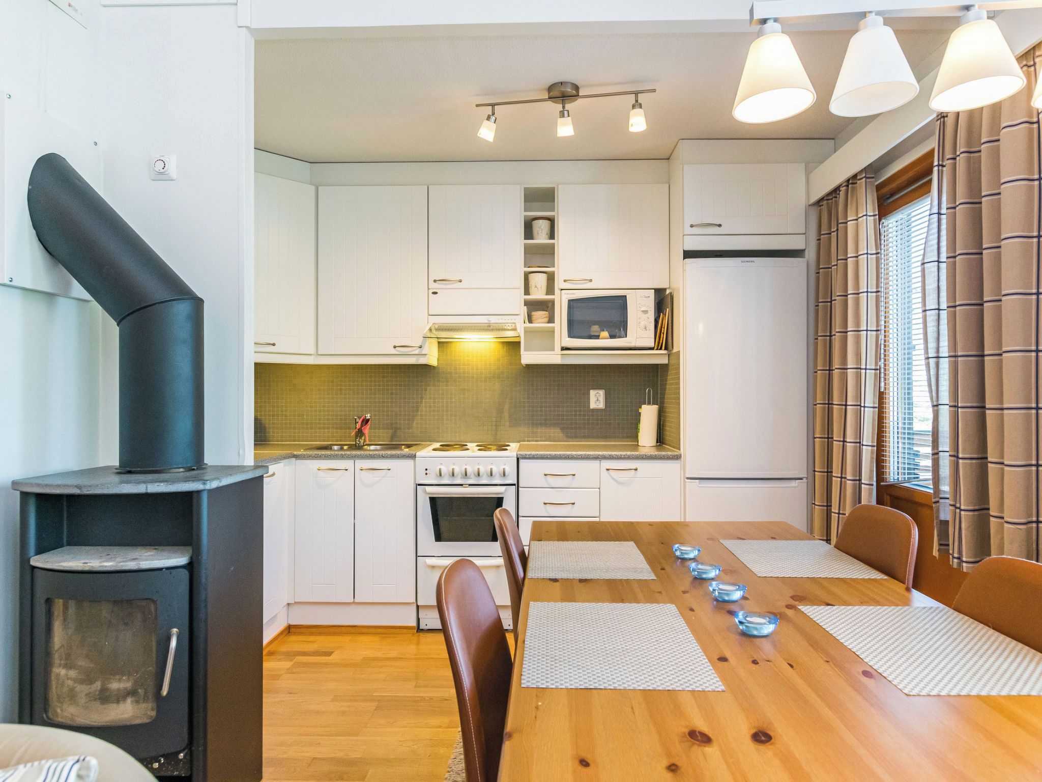A modern kitchen with white cabinets, a stove, oven, and a black wood stove. There is a dining table with chairs and a window with curtains.
