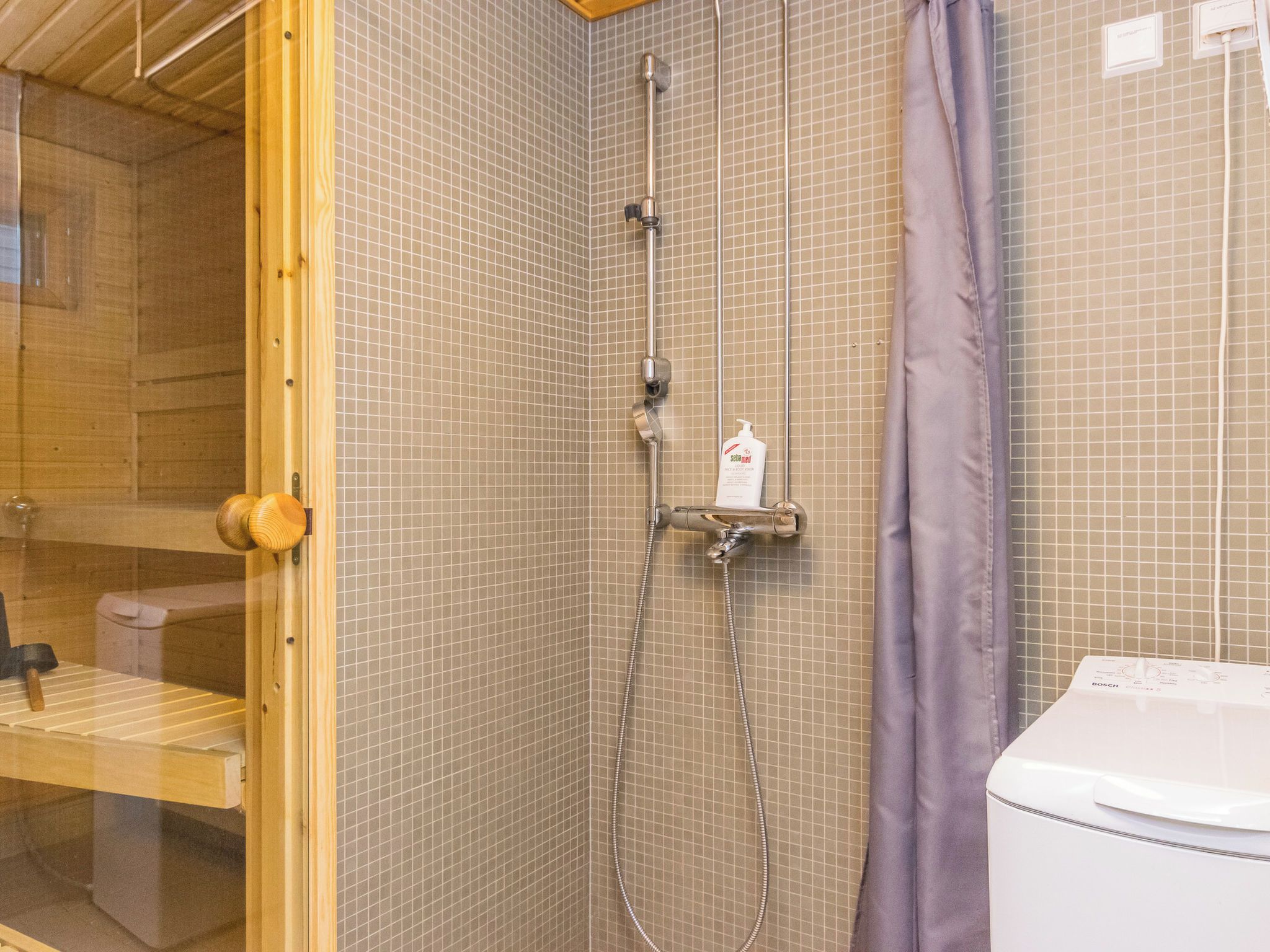 Suihkutilassa on käsisuihku, seinään asennettu hana ja pullo shampoota.
