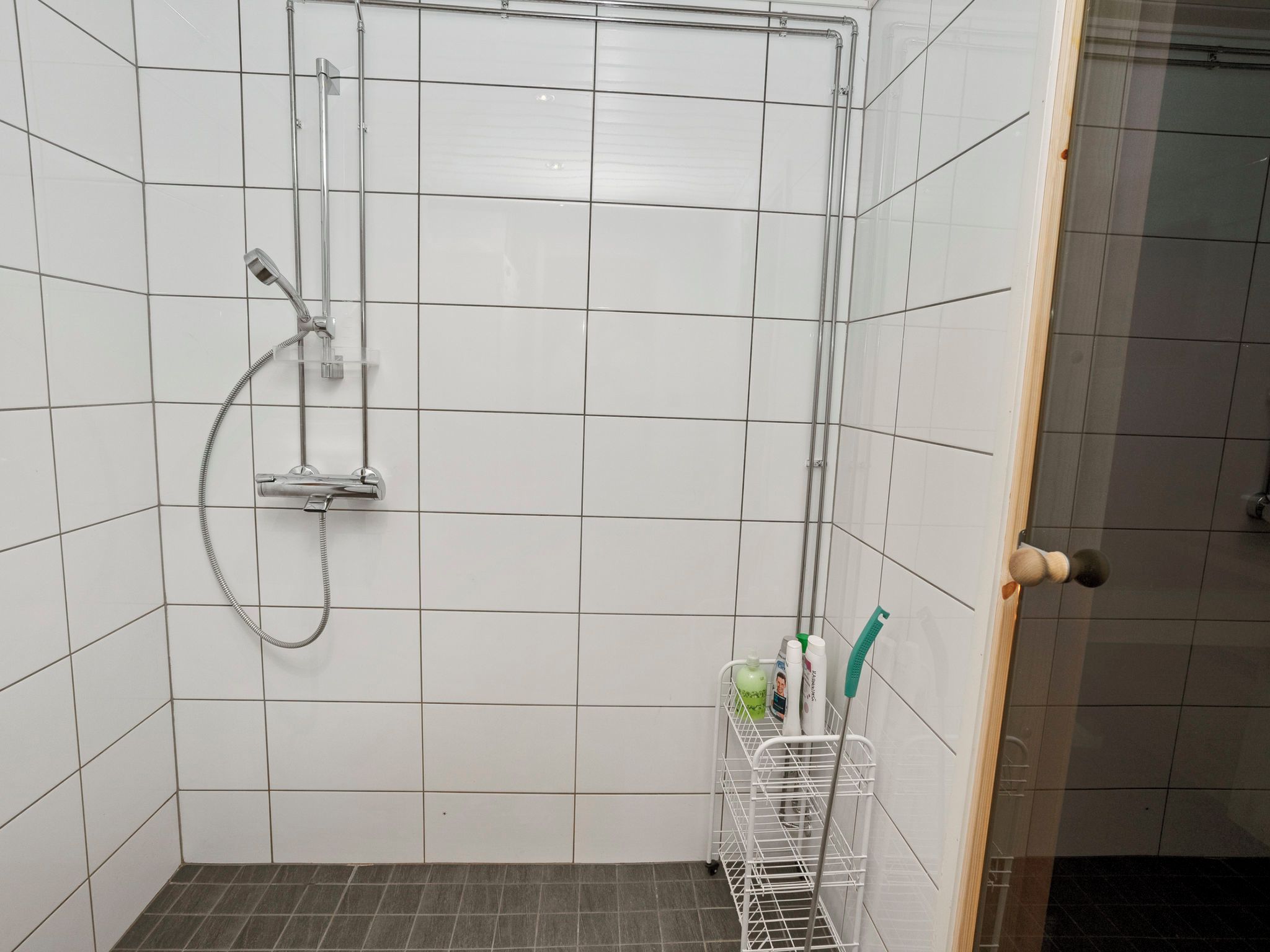 A small, tiled shower room with a showerhead, a hand shower, and a metal rack holding toiletries.