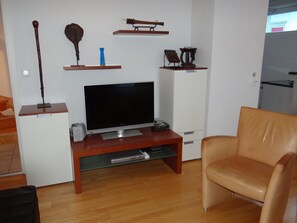 A living room with a flat-screen TV on a wooden stand, a leather armchair, and various decorative items on shelves.
