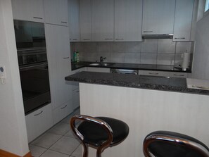 A compact kitchen with grey cabinets, a stainless steel sink, and a built-in oven. There are two bar stools with black seats.