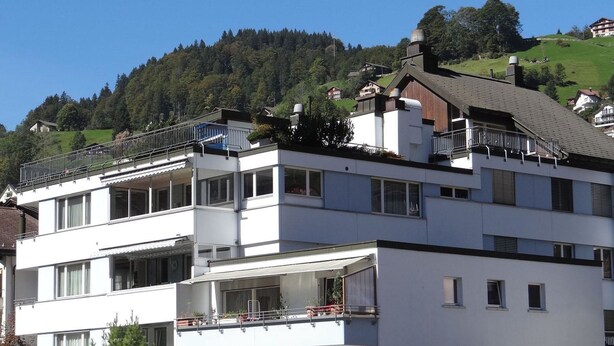 Ein mehrstöckiges Gebäude mit Balkonen, einer flachen Dachterrasse und Blick auf einen bewaldeten Hang.