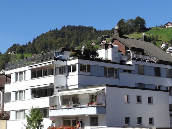 A multi-story building with balconies, a flat roof, and a view of a green hillside.