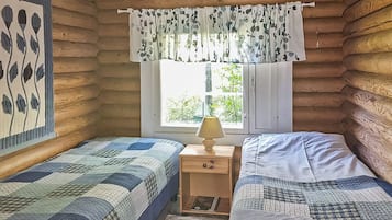 Mökkityyppinen huone, jossa on kaksi sänkyä, ikkuna, josta on näkymä, ja yöpöytä, jossa on lamppu.