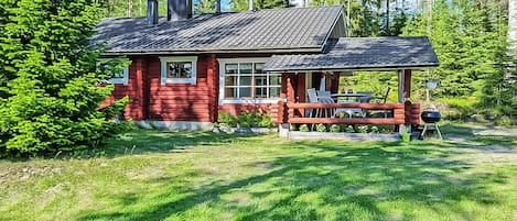 A red log cabin with a porch and outdoor seating, surrounded by lush greenery and trees.