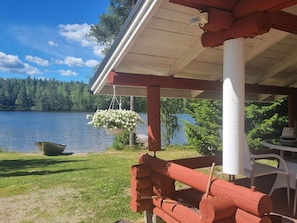 Eine Holzterrasse am Seeufer mit einer hängenden Blumenanordnung, einem Boot und Blick auf einen von Bäumen gesäumten See.