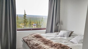A bedroom with a large bed, a view of trees and buildings, and a window with curtains. - Villa ylläs 203 by Interhome (Ylläsjärvi)
