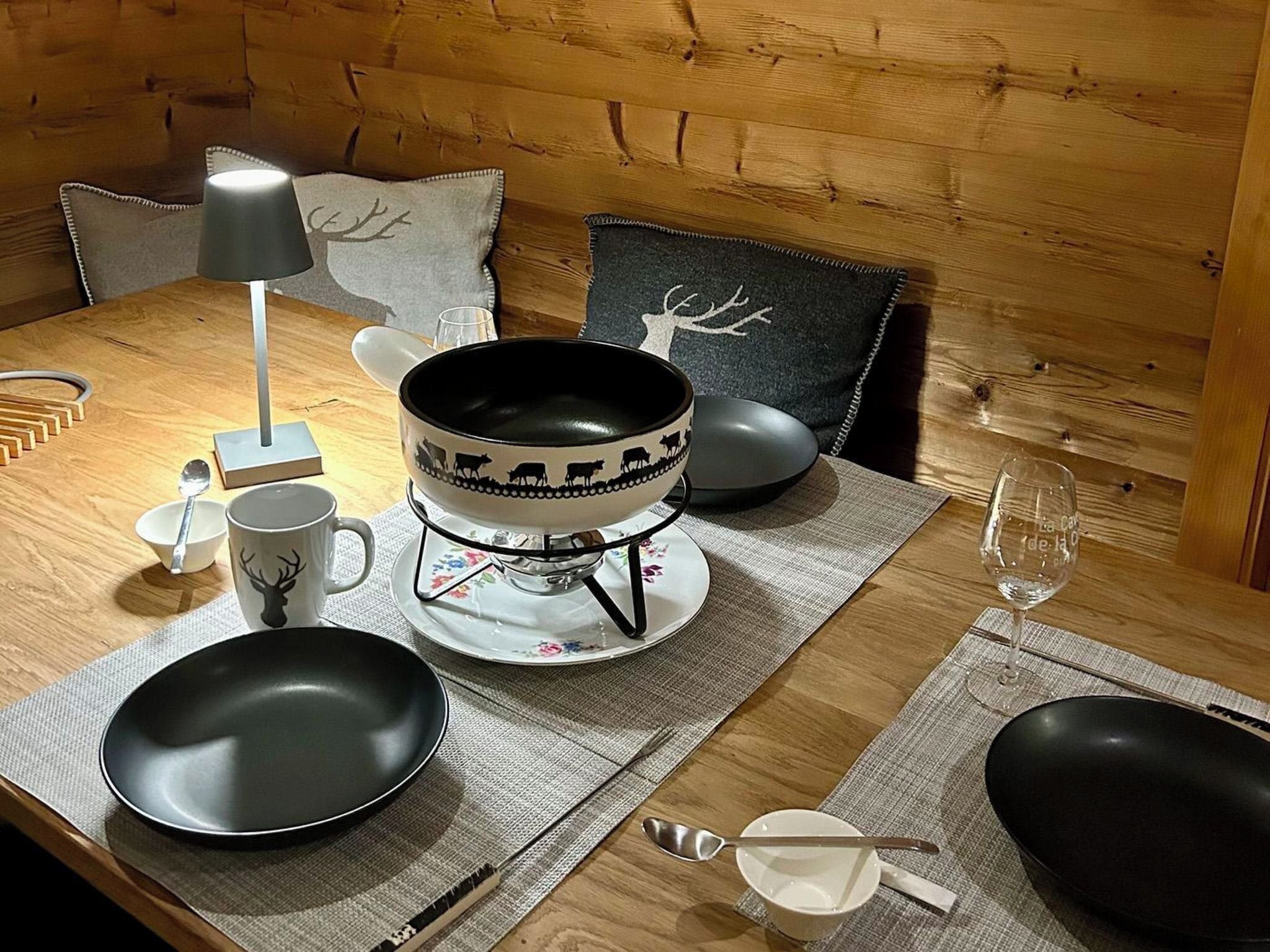 A wooden dining table with a black bowl, a white mug with a deer design, a glass, and a candle holder.